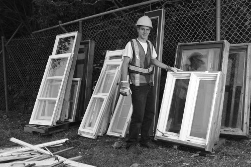A man wearing a safety helmet, high-visibility vest, and protective clothing stands outdoors beside a collection of window frames and sashes. The window frames are arranged in stacks, leaning against a wooden pallet on a grassy area. Behind him, there is a metal chain-link fence, and beyond that, some trees are visible. The frames are made of wood and glass, with some painted or finished surfaces, and vary in size and style. The scene likely depicts a waste clearance or rubbish removal for discarded or surplus windows, with the man from Commercial Waste Tring overseeing the collection. The environment appears to be an outdoor area or waste yard, suitable for rubbish disposal or recycling, with natural lighting indicating daylight hours. The overall scene reflects typical waste management practices, focusing on the removal of building or renovation debris such as unwanted window units, in a setting matching the context of rubbish removal services in the Tring area.