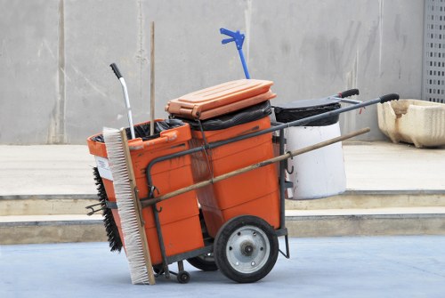 A mobile waste collection cart positioned on a paved outdoor area, used for rubbish removal services. The cart has a sturdy metal frame with two large black rubber wheels, and a bright orange plastic body designed to contain waste. On top of the cart, there is a flat, light brown plastic container or lid, and a blue plastic hook is attached to the side. Several long metal poles or handles extend from the cart, used for maneuvering or attaching equipment. A large white bin is situated to the right of the cart, and a building with a plain grey wall is in the background. The scene suggests a clean, organized environment, typical of commercial waste management activities, such as those provided by Commercial Waste Tring, serving nearby towns and postcode areas.