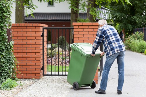 A man wearing a blue and white checked shirt and jeans is moving a large green wheeled rubbish bin towards a residential driveway in a suburban area. The bin is made of durable plastic with a textured surface and a closed lid. The driveway is paved with light-colored concrete or stone, leading up to a black metal gate supported by two red brick pillars. Behind the gate, there is a garden with various plants and trees, along with a house partially visible in the background. The scene is outdoors during daylight, with natural light illuminating the area and casting soft shadows from surrounding trees. The setting appears to be part of a waste collection or rubbish removal process handled by Commercial Waste Tring, serving the local community in the town or postcode implied by the context of the webpage about rubbish services.