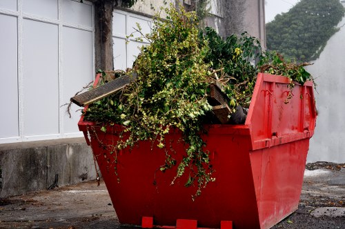 A large red plastic skip filled with garden waste, including green branches, leafy branches, and small twigs, positioned outdoors on a paved surface adjacent to a concrete wall. The skip's open top reveals a dense collection of organic debris. In the background, there is a white-panelled fence or wall with some trailing greenery and moss growing on it, indicating an outdoor environment. The lighting appears natural, casting soft shadows, and the overall scene suggests a waste removal service in a residential or commercial setting near Tring, with a focus on garden clearance or waste collection managed by Commercial Waste Tring. The scene captures the typical appearance of a rubbish collection point for organic or garden waste, relevant to local waste management services.