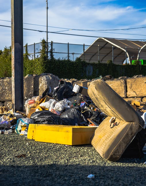A crumpled orange plastic rubbish bag lying on a textured asphalt pavement adjacent to a concrete curb and partially under a vehicle, with the top edge of a dark grey or black vehicle visible at the t