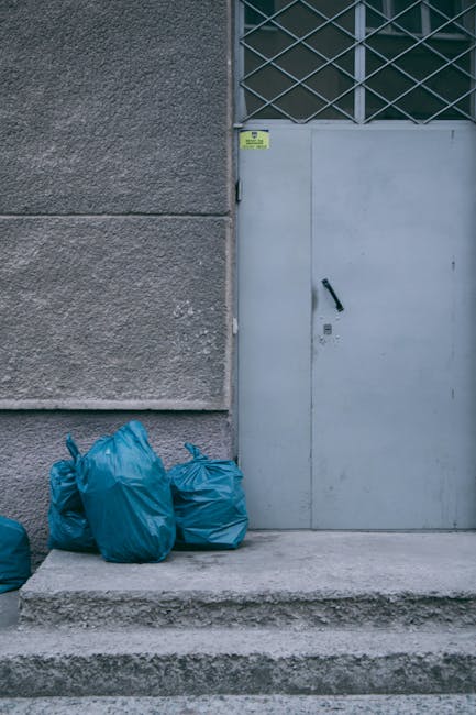 A roadside scene showing a large pile of mixed waste, including black garbage bags, a yellow plastic container, and a discarded, dirty car seat with a worn fabric cover, all resting on a gravel surfac