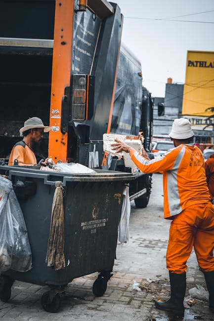 Two sanitation workers wearing high-visibility orange uniforms and white hats are engaged in rubbish collection outdoors on a paved street. One worker is seen tipping a large, rectangular, white conta