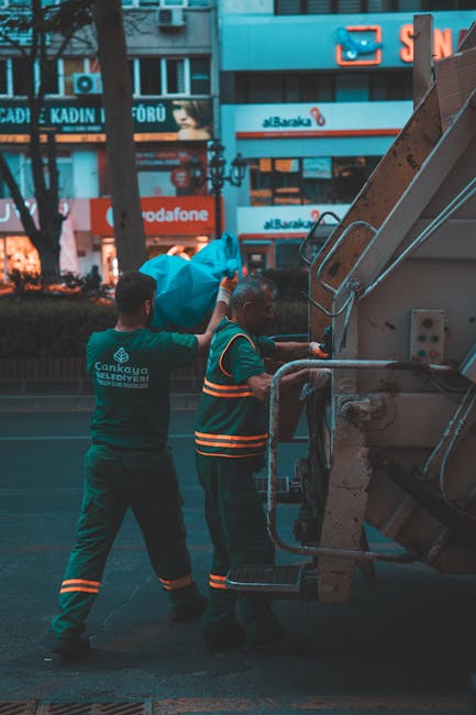 Two sanitation workers wearing high-visibility orange uniforms and white hats are engaged in rubbish collection outdoors on a paved street. One worker is seen tipping a large, rectangular, white conta